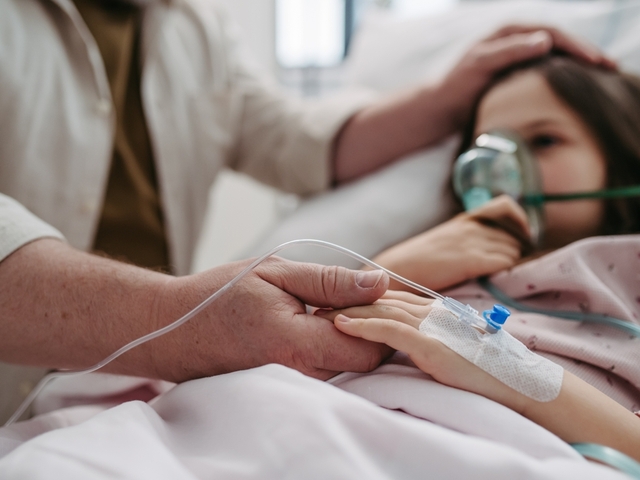 Ein Vater hält die Hand seiner Tochter. Das Kind liegt mit einer Sauerstoffmaske im Bett. An der Hand es Kindes ist ein intravenöser Zugang gelegt.