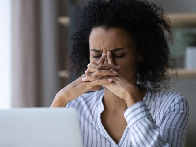Auf dem Bild ist eine Frau zu sehen. Sie hat dunkle Haare und trägt ein weiß-blau gestreiftes Hemd. Sie sitzt, vor ihr ist ein aufgeklappter Laptop. Ihre Hände sind ineinander verschränkt vor ihrem Gesicht, sie fasst sich mit ihren beiden Zeigefingern an die Nasenwurzel. Ihre Augen sind geschlossen, ihre Stirn ist gerunzelt. Sie sieht verzweifelt und angestrengt aus.   