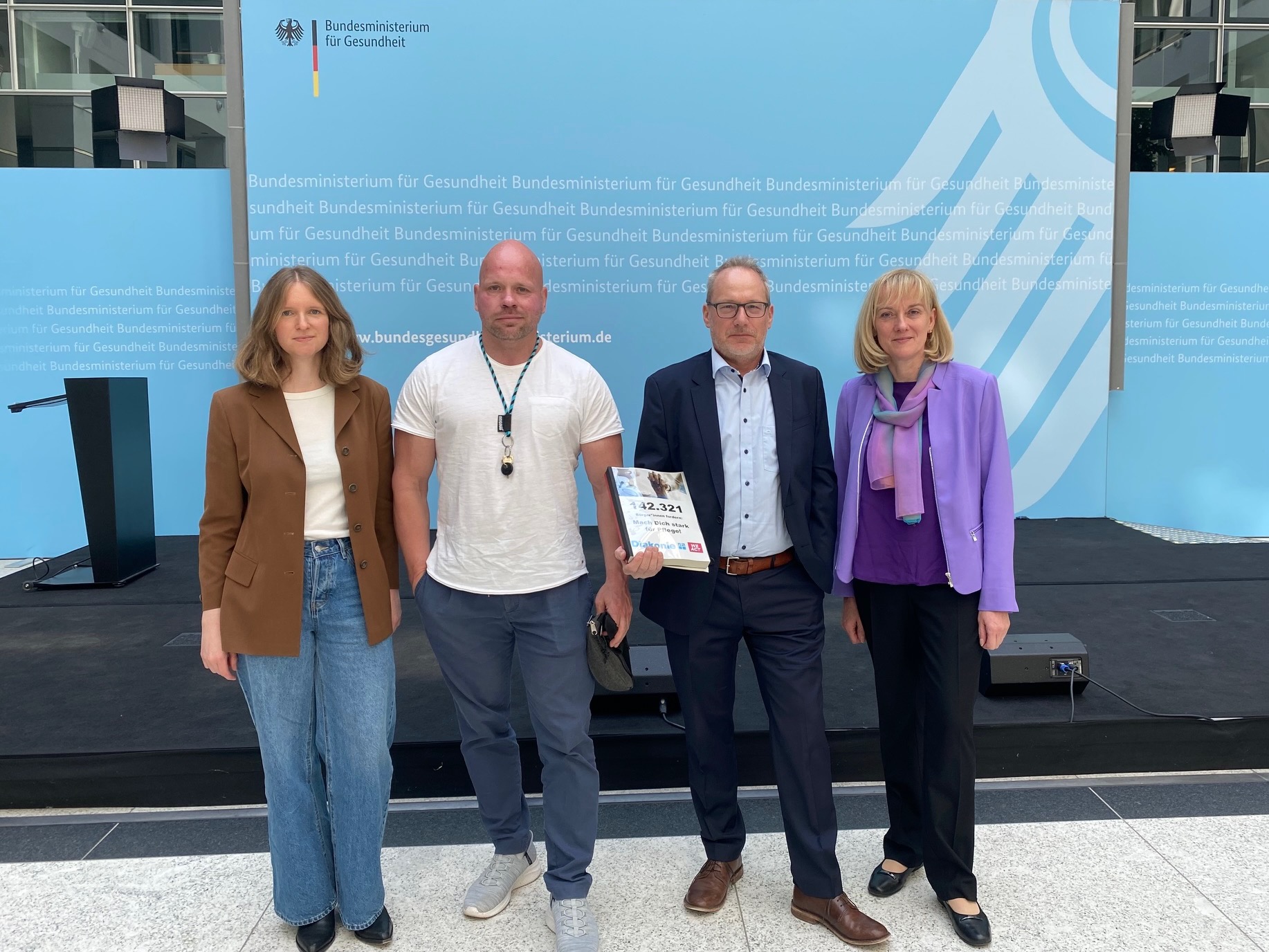 Auf dem Bild sind von links nach rechts zu sehen: Lisa Thelen, Pressereferentin von wir pflegen e.V., Ricardo Lange, Pfleger und Aktivist, Dr. Martin Schölkopf vom Bundesministerium für Gesundheit und Elke Ronneberger, Bundesvorstand Diakonie Deutschland. Herr Schölkopf hält ein Buch mit 143.000 Unterschriften für eine umfassende Pflegereform in der Hand.